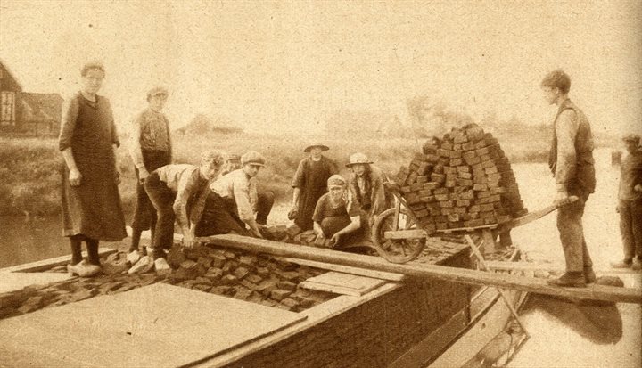 Een oude vergeelde foto uit 1927 laat een familie zien die turf opstapelt op een boot. Aan de rechterkant duwt een jongen een kruiwagen met een stapel turf over een loopplank van de kant naar de boot.